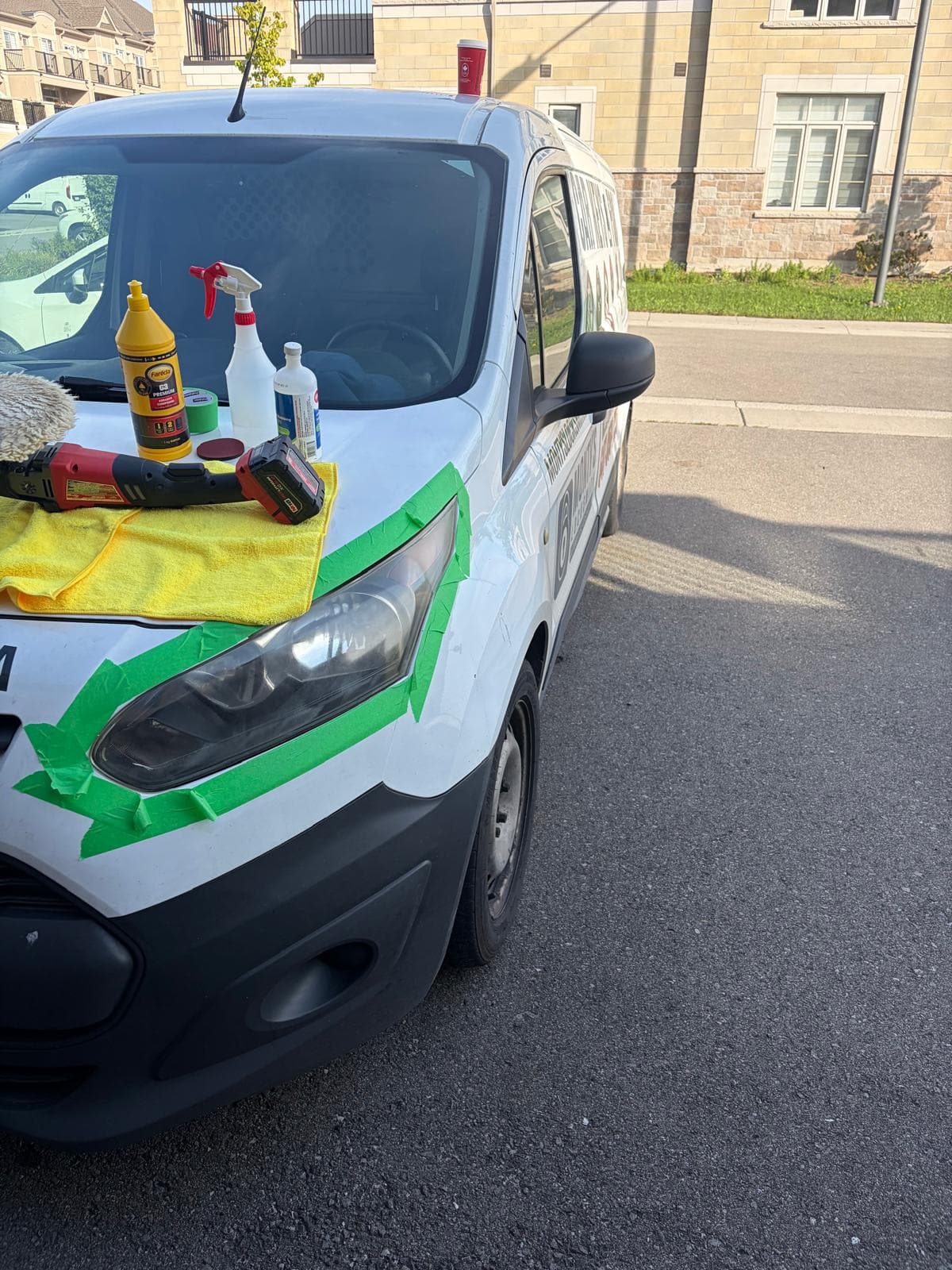 Machine polisher, compounds and masking tape laid out on a work van's hood, headlight taped off mid-job